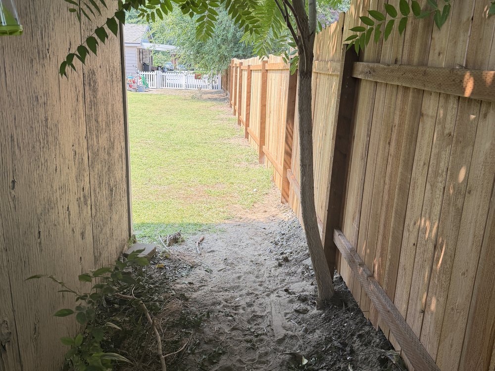 Narrow dirt gap between the cedar fence and a weathered plank wall, lawn and white picket fence beyond