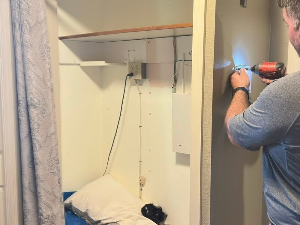 Open closet with a wood-edged shelf and bedding piled inside, someone using a drill on the wall beside it