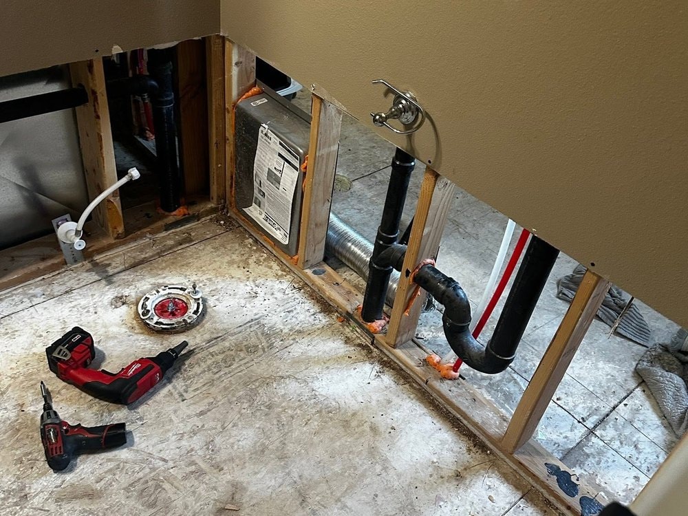 Bathroom stripped to bare subfloor with an exposed toilet flange, cordless tools and the wall cut open at the base