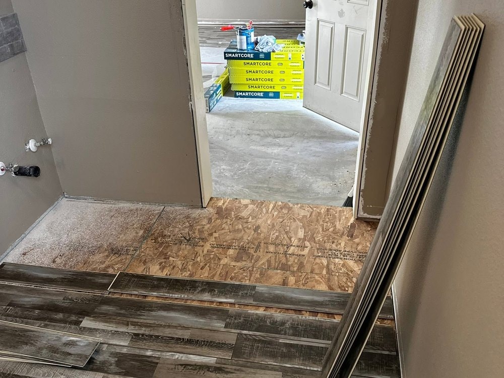 Vinyl plank flooring laid part way to a doorway, with bare subfloor and stacked flooring boxes beyond