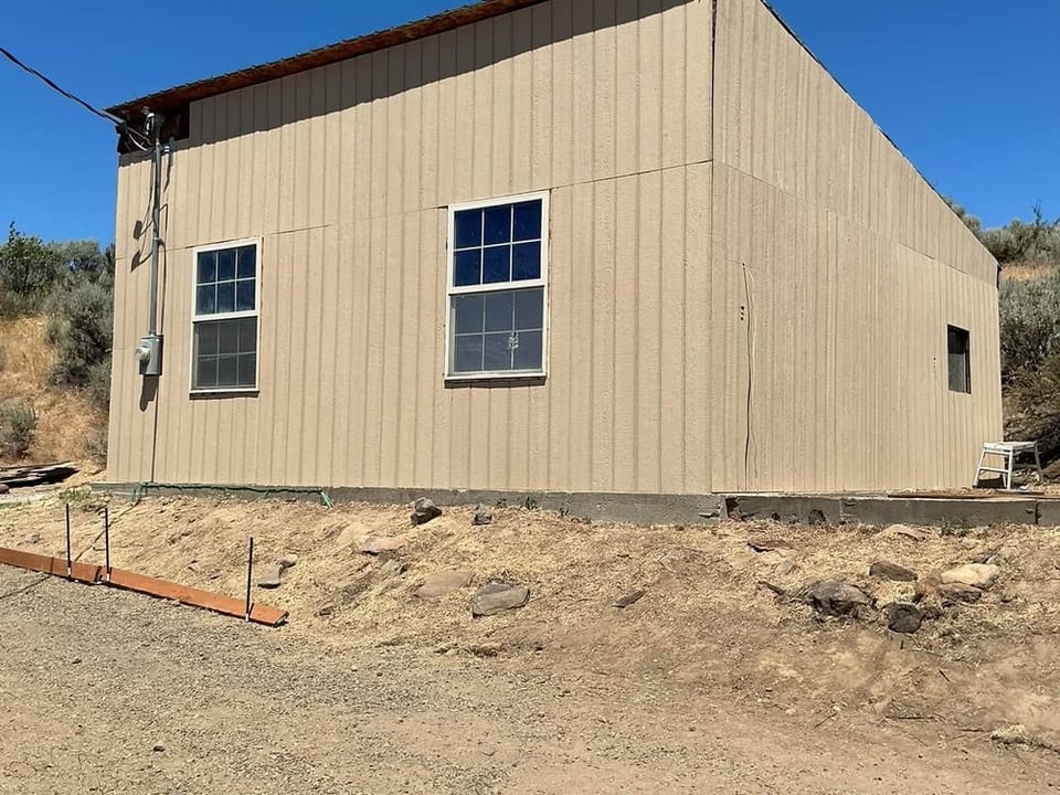 Detached rectangular building with tan vertical panel siding and two white framed windows on a dry sagebrush hillside.