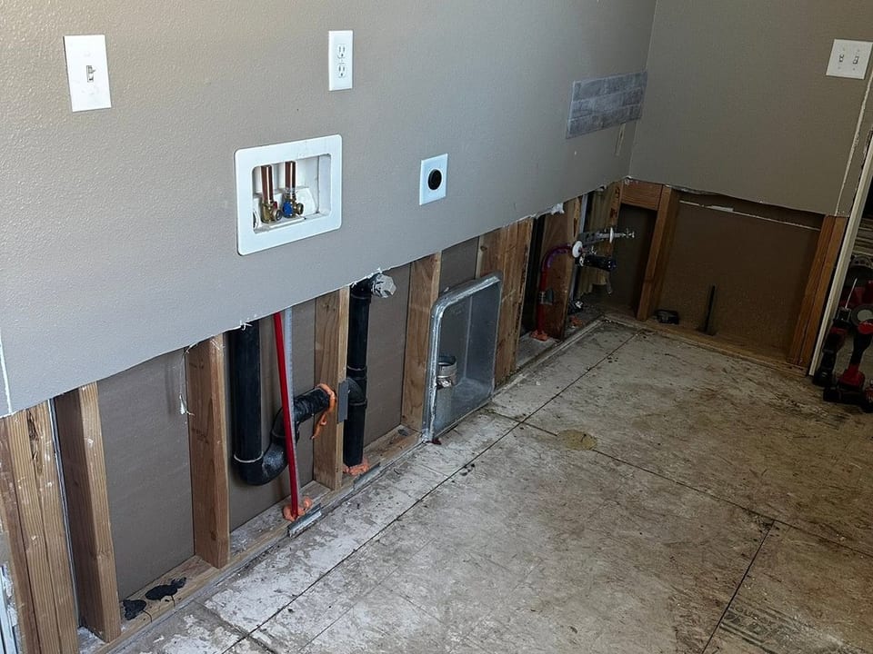 Interior wall with the lower drywall cut away exposing studs, plumbing lines and a dryer vent, flooring removed to subfloor.